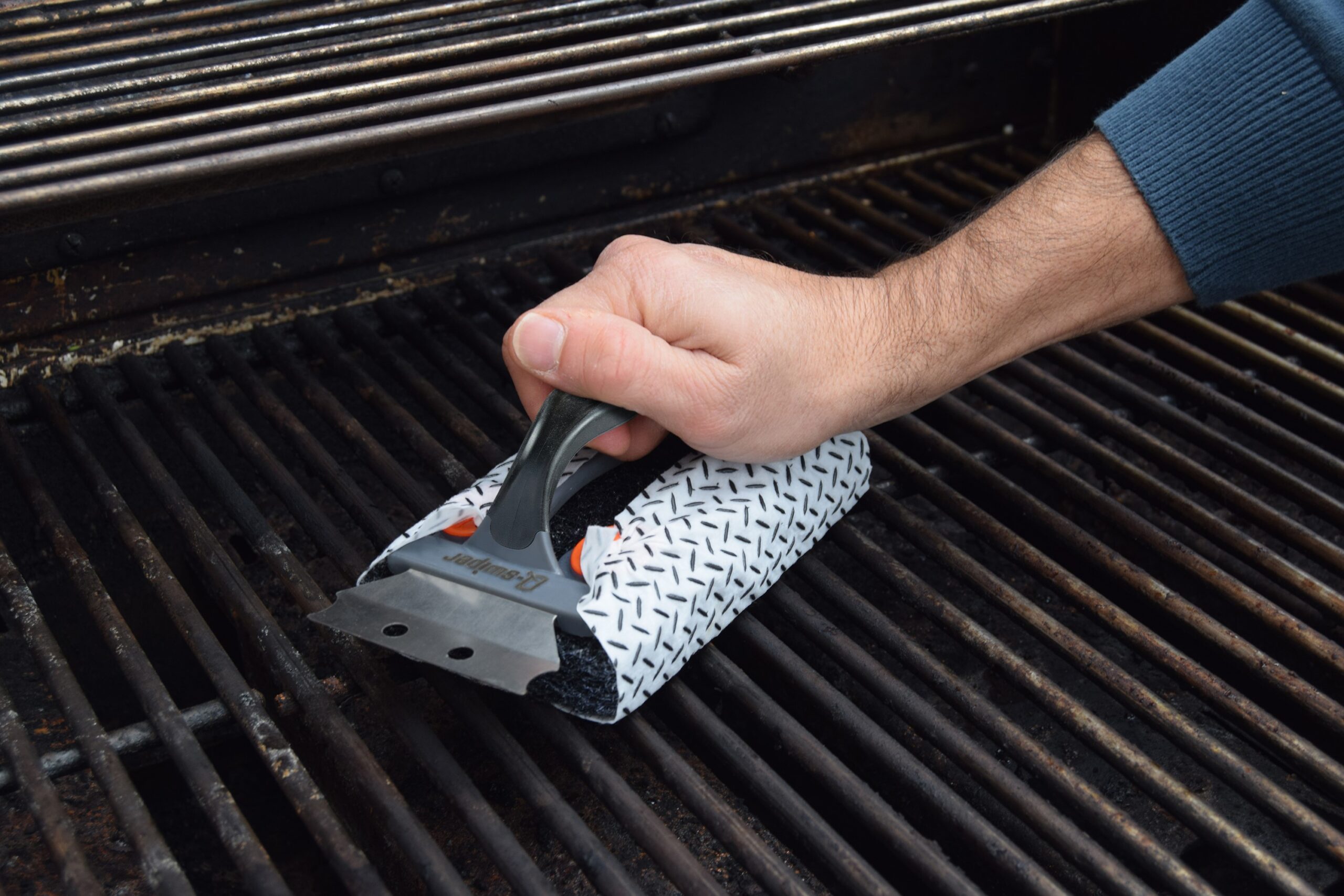 Close-up of a hand using the Q-Swiper bristle-free grill cleaning tool to scrub grease and residue off dirty grill grates